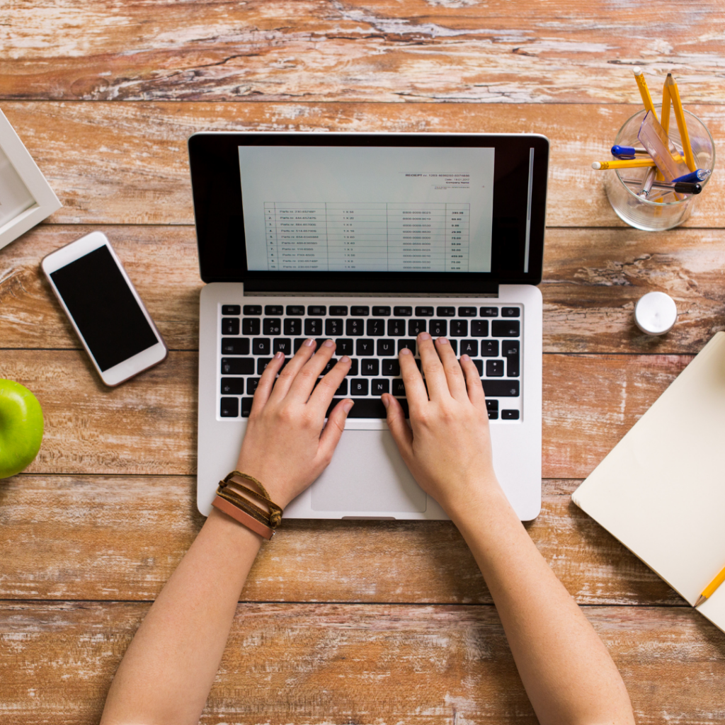 woman's hands hovering over laptop computer. iphone to the left, next to a green apple. to the right of the laptop, and open blank notebook. All on top of a wooden plank desktop.